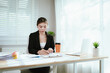 © Worawi - A young Asian businesswoman sits at her desk with a laptop and smartphone, smiling confidently while managing work in a modern office.