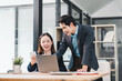 © PRIME STOCK LAB - Two Asian businesspeople, woman sitting and man standing, collaborate in modern office, smiling and discussing work at desk with laptop, smartphone, and documents