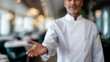 © Curioso.Photography - Smiling chef in uniform extending his hand in a welcoming gesture inside a fine dining restaurant.