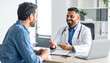 © amirhamzaaa - A doctor in a white coat smiles and gestures while consulting with a male patient at a desk in a bright office.