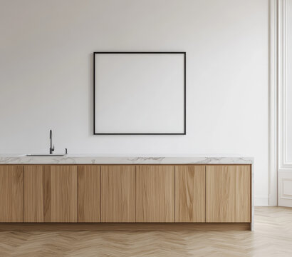 Minimalist kitchen interior with a large, square, black-framed artwork above a light wood and marble countertop, set against a white wall and herringbone wood floor
