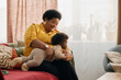 © Seventyfour - Happy African American grandmother holding little grandchild while playing with baby on cozy couch in living room