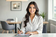 © f_bossa - A smiling young woman with long brown hair sits at a desk, writing with a pen while looking directly at the camera.