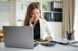 © wattana - Professional woman in office attire talking on the phone while working on financial tasks with a calculator and documents at her desk.