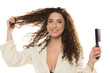© vladimirfloyd - Smiling woman with curly brown hair in white bathrobe holding hairbrush and touching hair against white background, happy hair care portrait