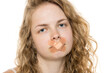 © vladimirfloyd - Young woman with curly blonde hair and band-aid over mouth, portraying censorship concept. Serious expression, close-up on white background.