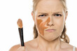 © vladimirfloyd - Young woman holding makeup brush with dissatisfied expression, displaying mismatched foundation streaks on face, showing frustration against white background