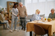 © Seventyfour - Elderly man using walker assisted by caregiver in care facility with other seniors in background. Room filled with books, chairs, and a table with breakfast items arranged
