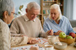 © Seventyfour - Elderly individuals engaging in board game with friends during daytime. Homey environment with bookshelves and fruits enhancing coziness and warmth in seniors' gathering space