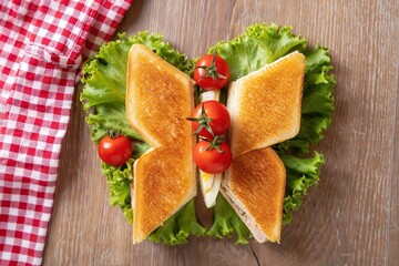 Poster - A sandwich cut into heart shape, with tomato and lettuce, on a wooden table covered by a red and white checkered cloth.