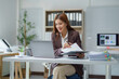 © Crystal - Young professional woman holding documents and using laptop while working at desk in modern office