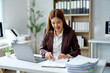 © Crystal - Young businesswoman smiling and reviewing financial documents and working on a laptop at her desk in a modern office