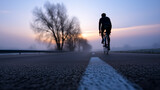 Cyclist riding on an empty road at dawn through misty countryside with the sun rising in the background.