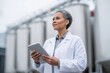 © Olha Havelia - Food safety inspector in a white coat documenting results on tablet at dairy production plant, steel tanks in background,