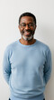 © Janos - Joyful middle aged dark skinned man in a light blue sweater and round glasses smiles brightly stands against a white wall looking relaxed and self sufficient.