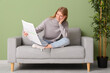 © Pixel-Shot - Beautiful young woman reading newspaper while sitting on sofa near green wall