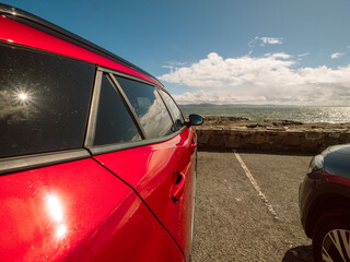 Wall Mural - A red car is parked in a parking lot next to a wall. The car is facing the ocean, and the sky is cloudy. Car park with stunning view on Galway bay and Burren mountains. Travel and tourism.