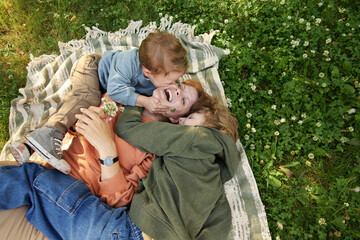 Wall Mural - Top view of two young boys kissing mother on cheek while laughing laying together on picnic blanket in green grass copy space