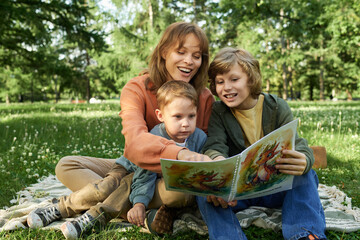 Wall Mural - Candid scene of mother and two boys laughing joyfully and pointing at pictures in storybook while sitting together on green grass in Summer park