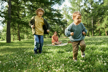 Wall Mural - Full length portrait of two young boys running towards camera on green grass while having fun in park with mother watching in background