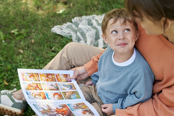 Wall Mural - High angle closeup of smiling young boy sitting on mothers lap and looking up playfully while reading book outdoors in green park copy space