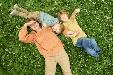 Wall Mural - Candid top view of happy mother lying with two sons on clover field and smiling up at camera playfully shielding faces from sunlight