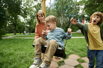 Wall Mural - Portrait of family with two children having fun at playground in park with happy little boy sitting in swing