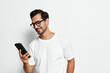 © SHOTPRIME STUDIO - Smiling man wearing glasses and a white t-shirt, looking at his mobile phone with joy, standing against a plain solid background. People lifestyle concept.
