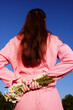 © SHOTPRIME STUDIO - A young woman holding flowers behind her back, wearing a pink outfit, with long brown hair, standing against a bright blue sky, evoking feelings of innocence and joy