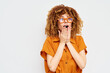 © SHOTPRIME STUDIO - Surprised young woman with curly hair wearing orange outfit and glasses, expressing astonishment against a plain light background Her hands cover her mouth in shock