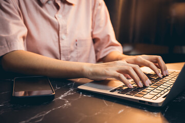 Poster - Close-up View of Hands Typing on Laptop with Smartphone on Table in Soft Warm Light for Productivity Concept