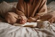 © Svitlana - Cozy scene: Woman sits cross-legged in bed writing in a notebook with a pen while enjoying coffee in a floral mug.
