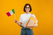 © Prostock-studio - Learning Italian, student exchange and travel. Happy lady holding notebooks and small flag of Italy, ready to study in college, standing over orange background, studio shot