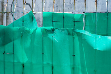 Poster - Fragmented green tarpaulin, secured to an aged metal fence with staples, presents a textured and weathered visual. The interplay of shadows emphasizes the layered effect and adds depth to the scene.