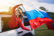 © Vladimir Razgulyaev - A woman joyfully waves the Russian flag from the window of a moving car as she embarks on a road trip to celebrate Russia Day. Dressed in a red outfit, she embodies the spirit of independence and