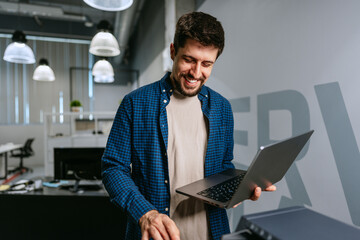 Poster - Man smiling while using a laptop in a modern office setting during a workday