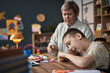 © DragonImages - Young boy with disability engaging in creative activity with help from teacher in classroom setting. Teacher supervising and assisting student with crafts at wooden table