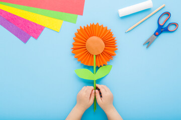 Wall Mural - Little child hands holding orange flower shape. Scissors, glue, pencil and colorful application paper on blue table background. Pastel color. Closeup. Creating gift. Point of view shot. Top down view.