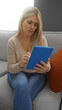 © Krakenimages.com - Woman sitting on sofa using a blue tablet in a cozy living room setting, creating a relaxed atmosphere with natural light and soft textures surrounding her.