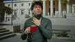 © Krakenimages.com - Man with coffee cup stands in front of roman columns at university campus holding his neck in discomfort expression on a sunny day.
