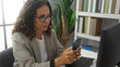 © Krakenimages.com - Woman in office using smartphone at desk, surrounded by computer, books, and plants, displaying a professional atmosphere in a modern, indoor work environment.