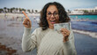 © Krakenimages.com - Middle-aged woman at seaside in uae holds a dirham banknote looking surprised and pointing, with a beach and water background showcasing a lively outdoor scene.