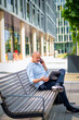© gzorgz - Middle aged man sitting on a bench in the city and using smart phone and notebook computer