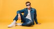 © Richafuji - A young man in denim jacket and jeans exhales a bubble while sitting against a solid yellow background. He wears sunglasses and white sneakers and socks, creating a casual fashion statement.