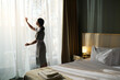 © pressmaster - Housekeeper in uniform drawing back curtains allowing sunlight to enter room revealing well-made bed with fresh towels on side table