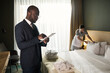 © pressmaster - Man in formal suit using tablet while hotel staff is arranging pillows on bed nearby creating a professional atmosphere