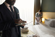 © pressmaster - Man in formal suit holding tablet in luxury hotel room while staff person making bed in background. Focus on professional setup and attentive service