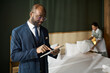 © pressmaster - Man in business suit using tablet in luxury hotel suite while young housekeeper is making bed in background