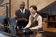 © pressmaster - Two professionals standing at reception desk discussing work details using technology and documentation. Man holding tablet while woman making notes at office desk