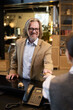 © pressmaster - Portrait of smiling elderly man with glasses holding credit card at hotel reception counter standing opposite staff. Background features office decorations and equipment